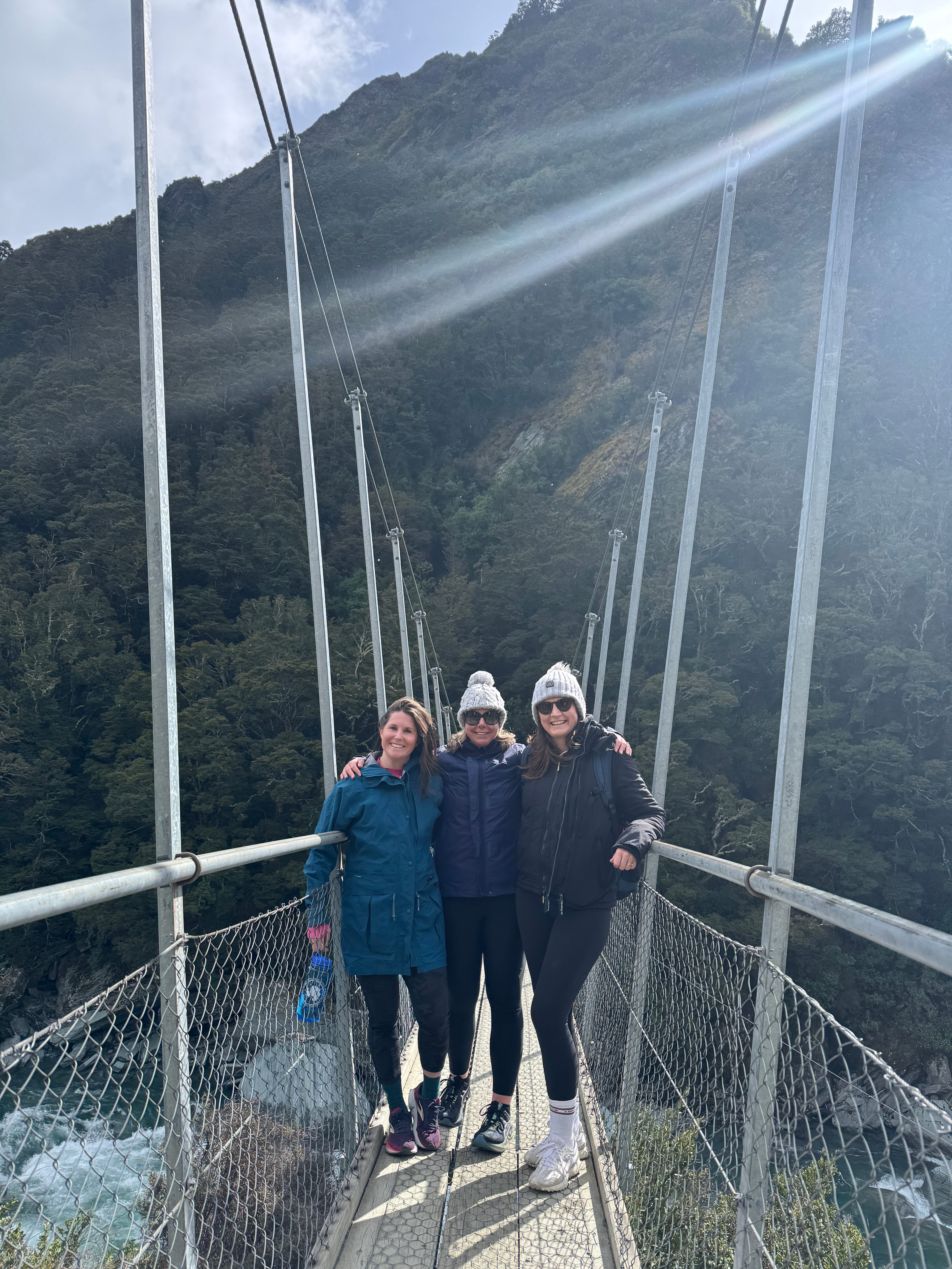 Three women on a suspension bridge surrounded by forested mountains
